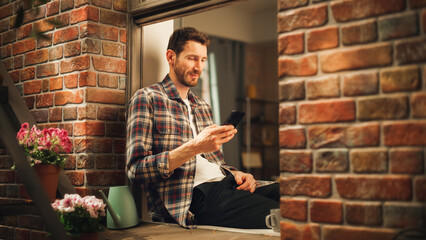 Leinwandbilder - Happy Handsome Caucasian Man Using Smartphone while Sitting on a Windowsill of His Cozy Apartment. Casual Shirt Wearing Guy Browsing Internet, Checking Videos on Social Media, Having Fun on Sunny Day.
