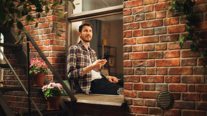 Leinwandbilder - Happy Handsome Caucasian Man Using Smartphone while Sitting on Windowsill of his Cozy Apartment. Casual Guy Wearing Shirt, Browsing Internet, Checking Videos on Social Media, Having Fun on Sunny Day.