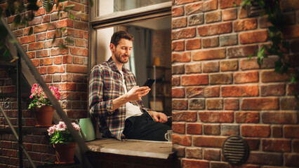 Leinwandbilder - Happy Handsome Caucasian Man Using Smartphone And Sitting on Windowsill of his Cozy Apartment. Casual Guy Wearing Shirt, Browsing Internet, Checking Videos on Social Media, Having Fun on Sunny Day.