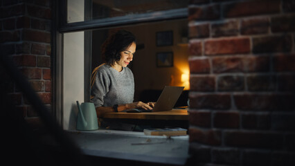 Leinwandbilder - Smiling Hispanic Businesswoman Sitting at Desk Working on Laptop Computer. Latin Female Entrepreneur Doing Virtual Remote Work from Home Office. Shot From Outside Into the Window of Apartment.