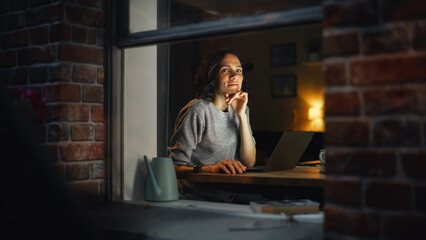 Leinwandbilder - Smiling Hispanic Woman at Home Using Laptop on the Warm Cozy Evening and Looking Thoughtfully. Professional Freelancer Working Remotely From Home. Shot From Outside Into the Window of Apartment.