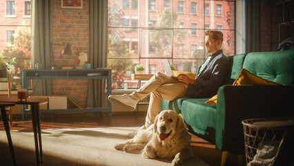 Poster - Handsome Young Man Using Laptop, Working from Home Living Room with Big Windows. Male Sitting on Couch, Using Computer, Golden Retriever Dog Sitting Next to Him and Wanting to Play or Go For a Walk.
