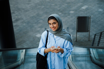 Wall Mural - Young muslim woman using cellphone while standing on escalator outdoors