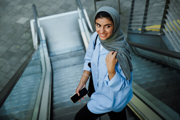 Wall Mural - Young muslim woman using cellphone while standing on escalator outdoors