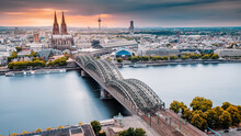 Cologne Aerial View With Trains Move On A Bridge Over The Rhine River On Which Cargo Barges And Passenger Ships Ply. Majestic Cologne Cathedral In The Background