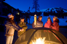 A Group Of Friends Stay Warm Outside At Night By Standing Next To A Firepit At A Ski Resort