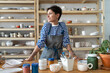 © DimaBerlin - Calm young female owner in pottery workshop looking at window and smiling, plans projects. Small business concept, ceramics studio. Successful italian woman entrepreneur with tattoos at workplace.