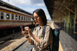 Traveler young asian woman using cellphone booking trip at terminal train station. Happy tourist travel by train using smartphone searching location. Female Backpacker arrival at platform railway.