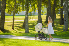 Young Adult Mother In Dress Pushing White Baby Stroller And Walking On Sidewalk At Town Park In Warm Sunny Summer Day. Spending Time With Infant And Breathing Fresh Air. Enjoying Stroll. Back View.