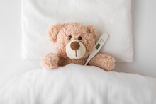 Teddy Bear With Digital Thermometer Lying Down On Pillow And Sheet Under Blanket In White Bed. Temperature Measuring In Cold And Flu Virus Time. Children Healthcare Concept. Closeup. Top Down View.