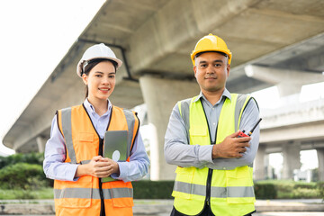 Wall Mural - Two Asian worker engineer man and woman architect looking construction with white safety helmet in construction site. Standing at highway concrete road site. Work planning with blueprint and tablet.