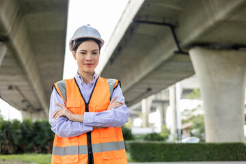 Wall Mural - Asian engineer worker woman or architect looking construction with white safety helmet in construction site. Standing at highway concrete road site. Progress planning of highway bridge.