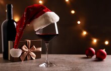 Glass Of Red Wine With Santa Claus Hat, Bronze Gift Box And Christmas Decorations On Table. Christmas Lights In The Black Background.