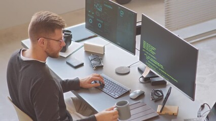 High angle shot of Caucasian male IT specialist looking at data and program codes on two computer monitors while working on new software or application in office