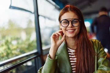 Beautiful woman with red hair and glasses sits in a cafe in the city and looks out the window smile with teeth, female freelance blogger close-up