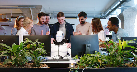 Robot sitting at table with computer surrounded by diverse people team of coworkers. Humanoid employee showing thumbs-up. Males and females developers with cyborg in office. Artificial intelligence