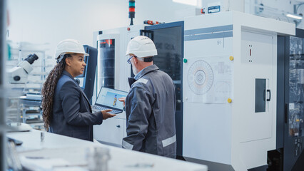 Two Diverse Professional Heavy Industry Engineers Wearing Safety Uniform and Hard Hats Working on Laptop Computer. African American Technician and Worker Talking Next to a Factory Machine.