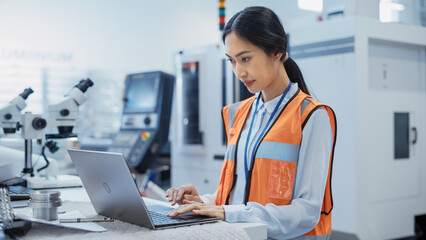 Fototapete - Portrait of an Asian Female Engineer in Orange Safety Vest Working on Laptop Computer at Electronic Manufacturing Factory. Technician Working on Daily Tasks and Research and Development Data.