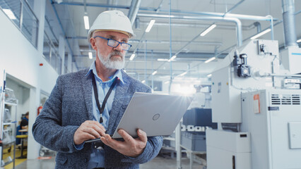 Leinwandbilder - Portrait of a Bearded Middle Aged General Manager Walking in a Factory Facility, Wearing Casual Suit and a White Hard Hat. Heavy Industry Specialist Working on Laptop Computer.