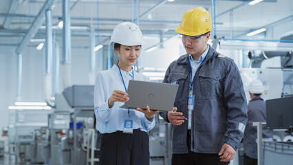 Wall Mural - Portrait of Two Young Asian Heavy Industry Engineers in Hard Hats Standing with Laptop Computer and Discussing Work Process in a Factory. Two Manufacturing Employees Chatting in Production Facility.