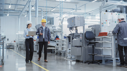 Leinwandbilder - Wide Shot: Two Heavy Industry Employees in Hard Hats Discussing Job Assignments at the Factory, Using Laptop Computer. Asian Engineer and Technician at Work Smiling