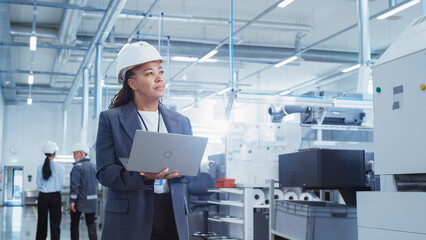 Portrait of a Black Female Engineer in Hard Hat Standing and Using Laptop Computer at Electronic Manufacturing Factory. Technician Thinking About Daily Tasks and Working on Project Pipeline.