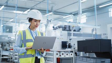 Fototapete - A Focused and Professional Asian Female Engineer in White Hard Hat Standing with Laptop Computer at Electronic Manufacturing Factory. Successful Employee Working on Daily Tasks