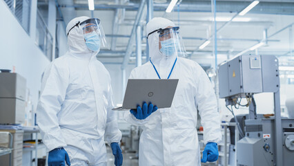 Wall Mural - Two Scientists Walking in a Heavy Industry Factory in Sterile Coveralls and Face Masks, Using Laptop Computer. Examining Industrial Machine Settings and Configuring Production Functionality.