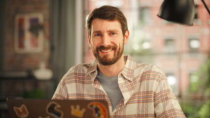 Poster - Portrait of a Handsome Bearded Young Adult Man Using Laptop Computer with Stickers on the Back in Creative Loft Living Room at Home. Happy Specialist in Checkered Shirt Looking at Camera, Smiling.
