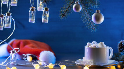 Wall Mural - A cup of cocoa or hot chocolate with marshmallows on the Christmas table. Santa hat, garlands and Christmas tree branches, copy space