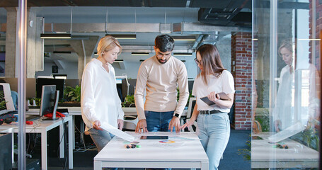 Working team of man and two women standing over the table with tablet device and charts and discussing project. IT employees. Mixed-races male and females talking and brainstorming. Startup concept.