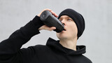 Fototapeta Młodzieżowe - Male runner drinking water from a black plastic bottle after a cardio workout. Sportsman in black hoodie and cap hydrating during outdoor training.