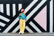 © okrasiuk - Hipster fashion young woman in bright clothes, sun glasses and bucket hat posing on the painted brick wall background. Urban city street fashion. Selective focus. Copy space