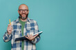 © Iona - Cheerful Caucasian bald beardy man in plaid shirt holds book and pen looks at camera against turquoise  studio backdrop. Excited Italian male in glasses, teacher has idea. Mockup, education, knowledge