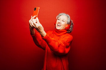 Senior woman wearing red clothes, taking a selfie with red headphones and red smartphone, over a red background