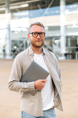 Vertical photo of a positive smart confident bearded caucasian man, with glasses, in a casual clothes, a programmer, seo, holds a laptop in his hand, looks at the camera, smiles friendly