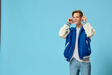 Wall Mural - cute, handsome guy in a bomber jacket listens to music in big headphones holding them with his hands. Studio photography on a blue background with space for an advertising mockup