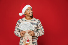 Merry Fun Young Man Wear Sweater Santa Hat Posing Hold Stocking Sock Fan Of Cash Money In Dollar Banknotes Look Aside Isolated On Plain Red Background. Happy New Year 2023 Celebration Holiday Concept.