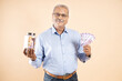 © GAJENDRRA BHATI  - Happy senior Indian man holding jar full of money and notes of 100 rupee isolated on beige background, Asian mature male with lot of cash in hand.