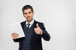 © LIGHTFIELD STUDIOS - cheerful young businessman in suit holding digital tablet and showing thumb up isolated on grey