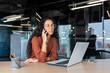 © Liubomir - Young beautiful businesswoman talking on the phone and looking towards the window, happy hispanic woman inside the office using a laptop at work
