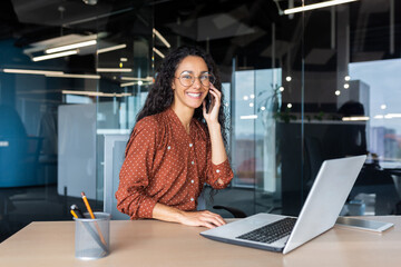 Wall Mural - Portrait of successful latin american business woman, office worker smiling and talking on phone looking at camera inside office, accountant financier using laptop at work.