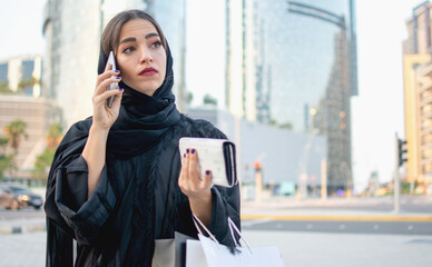 Wall Mural - Beautiful young muslim woman in traditional clothes holding shopping bags and wallet and talking on mobile phone on the street.