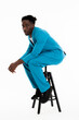© ABCreative - Side of the view young african american man model sitting on black chair on white background in studio isolated posing fashionably wearing blue casual suit lifstyle concept copy space.