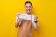 © Bangun Stock Photo - Excited young Asian man 20s barista employee in brown apron working in coffee shop, holding a gift store coupon voucher card, celebrating success on yellow background. Small business startup concept