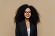 Portrait of beautiful Afro American woman with crisp hair, dressed in elegant black jacket, transparent glasses, looks directly at camera with gentle smile wears optical glasses isolated on brown wall