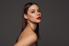 Portrait Of Female Fashion Model With Naked Shoulders And Long Hair Looking At Camera Over Dark Grey Background. Beautiful Woman With Trendy Make-up, Red Lipstick And Well-kept Skin.