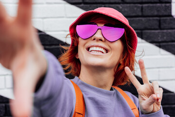 Wall Mural - Close-up excited redhead woman in magenta heart-shaped sunglasses smiling and showing V finger sign while taking a selfie photo outdoors. Color of the 2023 year. Emotional hipster fashion women