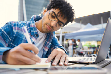 Wall Mural - Young pensive middle eastern freelancer using laptop computer, taking notes, working online at workplace. Handsome curly haired student studying, exam preparation. Online education concept 