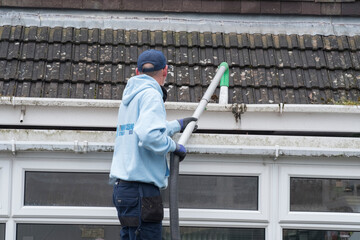 A window and gutter cleaner cleaning Dirty clogged white plastic pvc gutters and drain pipes with mossy green mould on plastic fascias. Blocked drains and guttering need  regular yard work maintenance